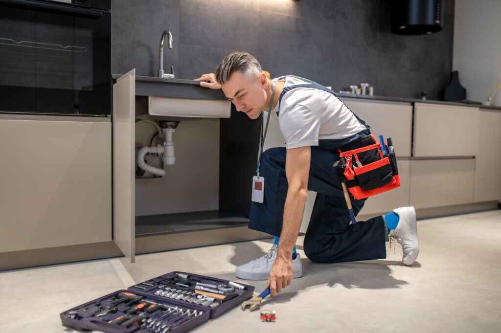 man-crouched-near-sink-stretching-out-hand-wrench